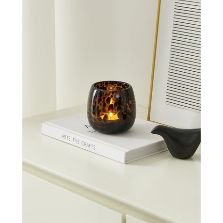 Mandav Hurricane Lantern, a rounded glass candle holder in tortoiseshell amber and black, lit and placed on a white book beside a framed print on a shelf.