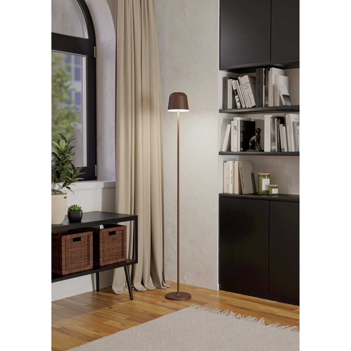 Mannera-L LED Floor Lamp in auburn finish with slim stem, bell-shaped head and round base, shown lit beside a curtained window in a living room with bookshelves.