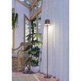Mannera-L LED floor lamp in auburn finish with bell-shaped head and round disc base, shown lit beside sheer curtains in a plant-filled conservatory setting.