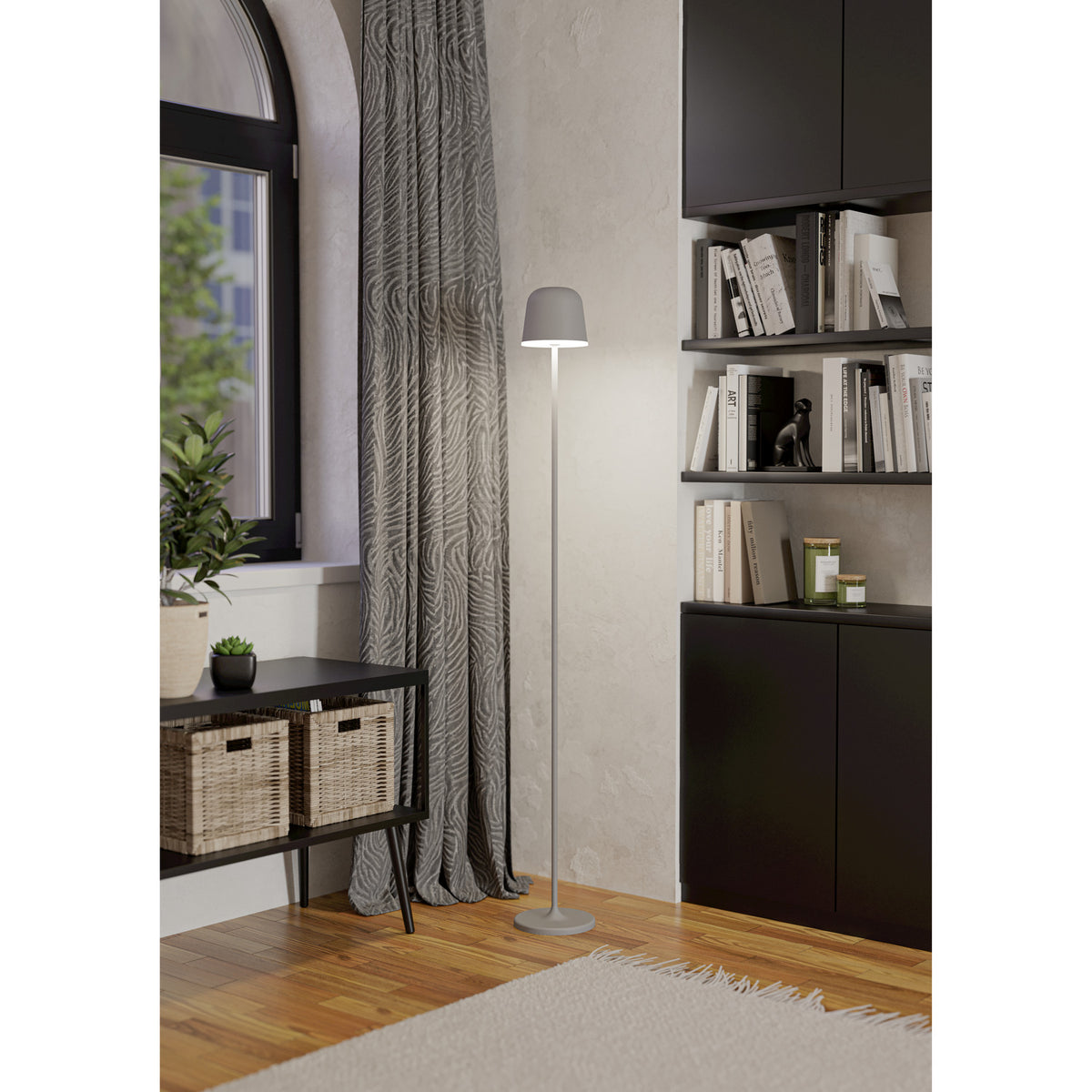 Mannera-L LED floor lamp in grey, with a slender stem, dome shade and round disc base, shown lit beside a curtained window and black bookshelf in a living room.