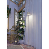 Mannera-L LED floor lamp in sand finish with bell-shaped shade on a slim stem and round base, shown lit beside sheer curtains in a plant-filled living space.
