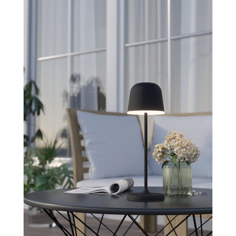 Mannera black LED touch table lamp with rounded dome head and slim stem, lit on an outdoor coffee table beside a flower vase and sofa.