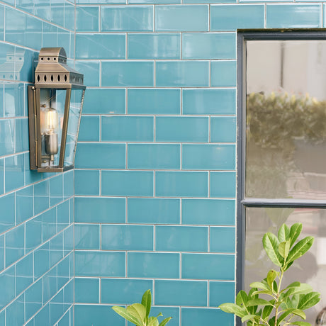 A wall adorned with gleaming teal subway tiles features the Mansion 1 Light Outdoor Wall Light in aged brass, adding a touch of elegance with its handcrafted design. A window is partially visible on the right, while lush green plants thrive in the bottom foreground, enhancing the scene with a hint of nature's charm.