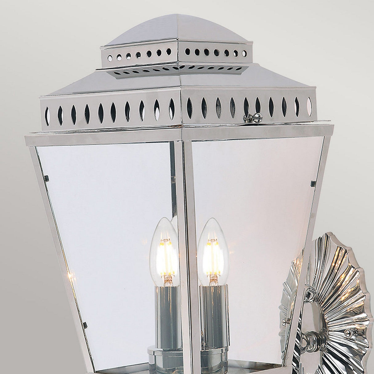 A close-up of the Mansion 3 Light Outdoor Wall Light highlights its polished nickel finish and lantern-inspired design. It features glass panels that reveal the three light bulbs inside, topped with decorative vent holes and mounted on a detailed metal base.