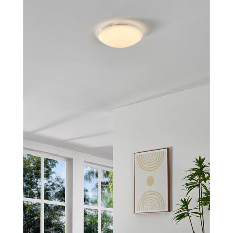 Margitta 1 LED flush ceiling light in white, round frosted glass diffuser with swirling line pattern, shown illuminated on a hallway ceiling near a framed print and plant.