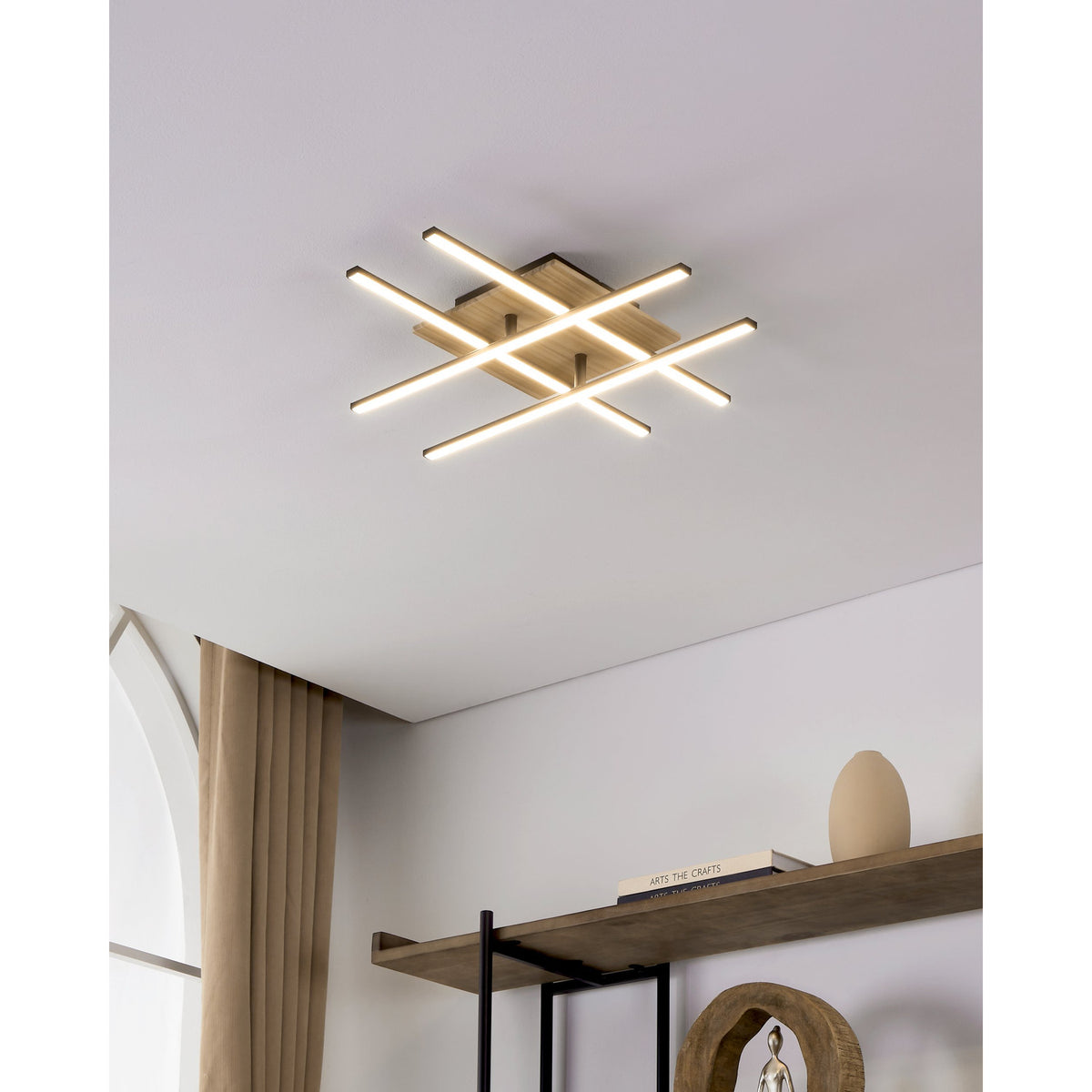 Marinello 1 LED flush ceiling light with four black bars crossed over a square wood plate, illuminated in warm white on a white ceiling above a shelf styled with books and decor.