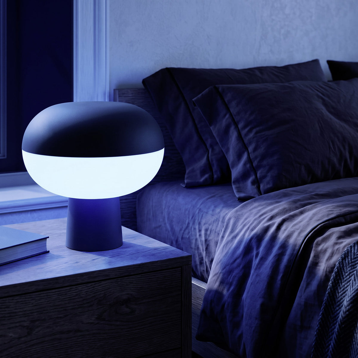 Marini Smart Colour Changing LED Table Lamp in matt black with glowing white shade band, shown lit on a dark bedside table beside a bed in a dim bedroom.