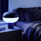 Marini Smart Colour Changing LED Table Lamp in matt black with glowing white shade band, shown lit on a dark bedside table beside a bed in a dim bedroom.