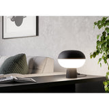 Marini Smart Colour Changing LED Table Lamp in matt black, with rounded mushroom-shaped shade illuminated white, shown on a dark console table in a living room setting.
