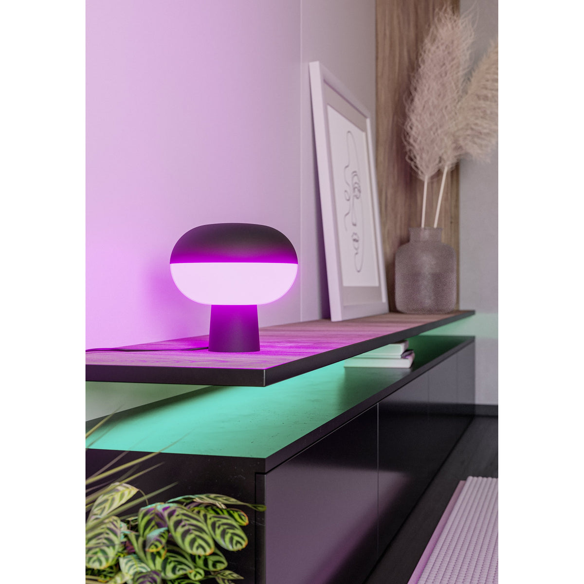 Marini Smart Colour Changing LED Table Lamp in matt black, mushroom-shaped with glowing pink band, shown on a wooden shelf beside a framed print and vase in a living room setting.