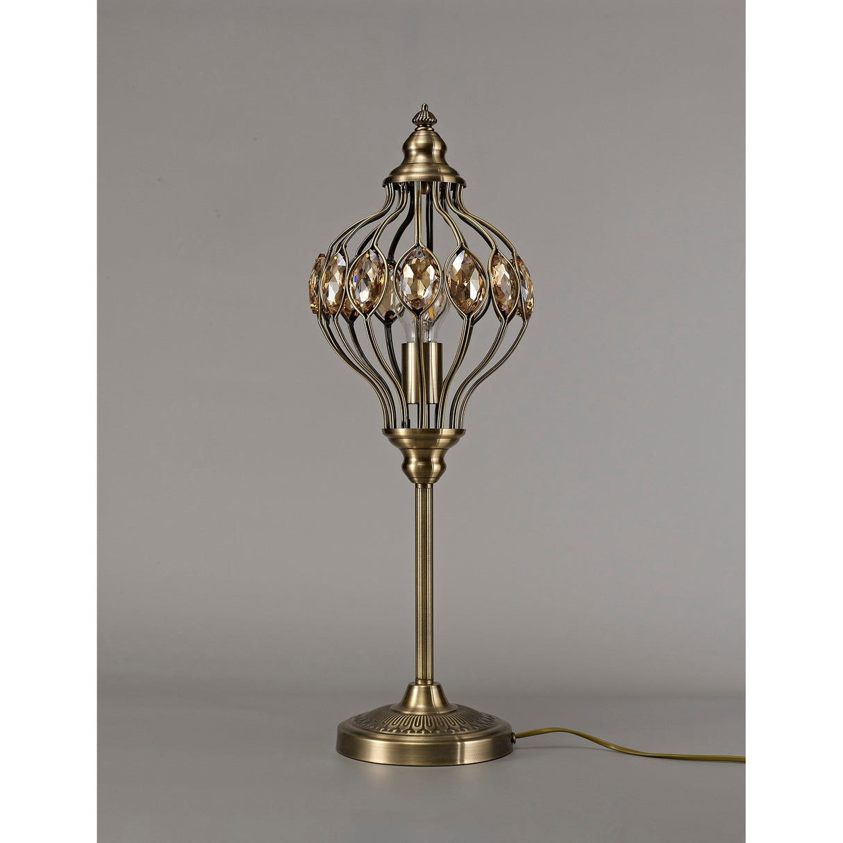 The Marisa 1 Light Crystal Table Lamp features an antique brass finish and amber crystal accents, with a cage-style shade inspired by Middle Eastern lanterns. It stands on a round base with a yellow cord against a plain gray background.