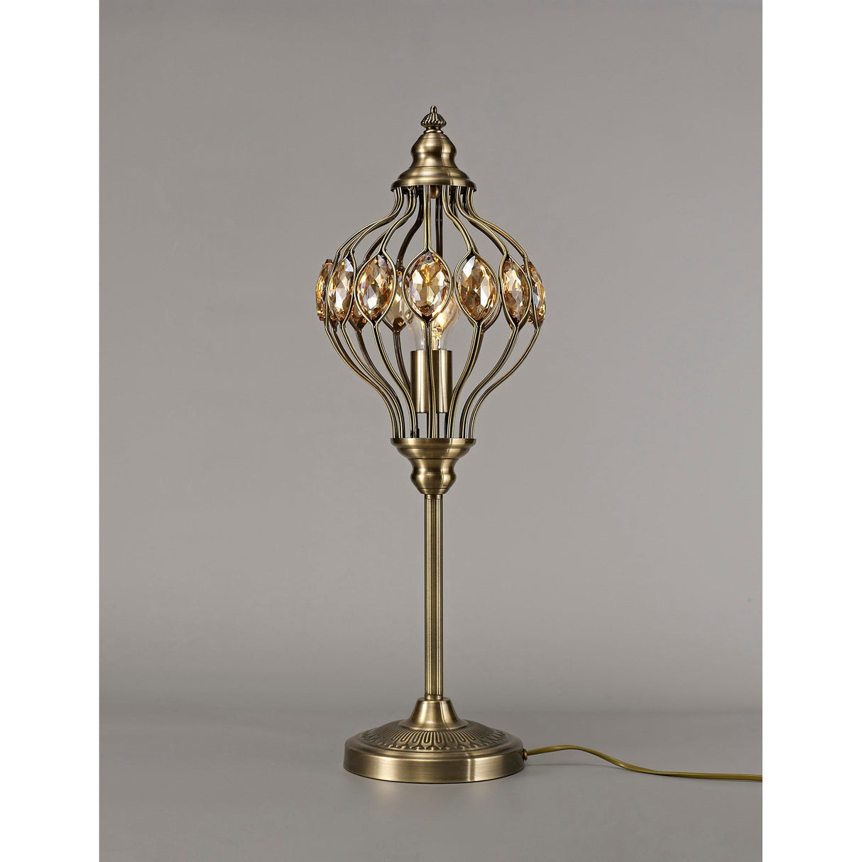 The Marisa 1 Light Crystal Table Lamp in antique brass with amber crystal accents stands on a plain surface, showcasing its vintage-inspired openwork metal shade and round base against a neutral gray background.