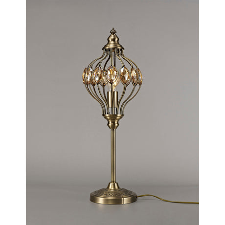 The Marisa 1 Light Crystal Table Lamp in antique brass with amber crystal accents stands on a plain surface, showcasing its vintage-inspired openwork metal shade and round base against a neutral gray background.