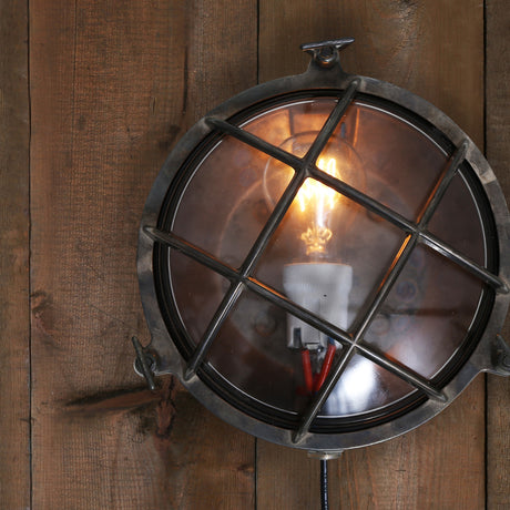 Marlune Small Bulkhead Wall Light IP54 in aged silver-toned metal, round glass front with diagonal cage grille and lit bulb, mounted on a wooden plank wall.