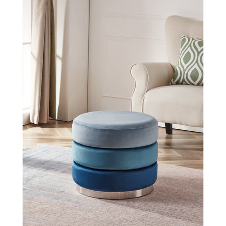 Mashike Round Pouffe in Blue, three stacked upholstered tiers graded pale sky blue to navy on a silver-toned metal base, shown beside an armchair in a living room.