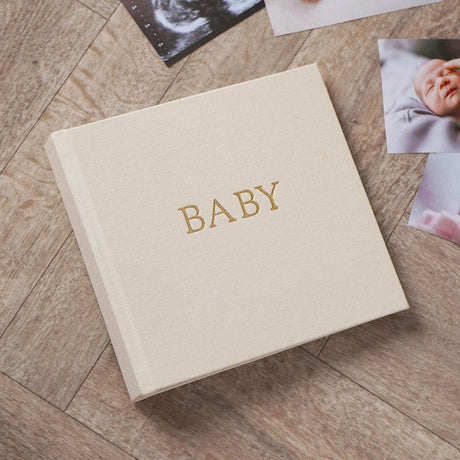 Square baby photo album in cream linen with gold-embossed "BABY" text, shown on a wooden floor beside scattered baby photographs and an ultrasound print.