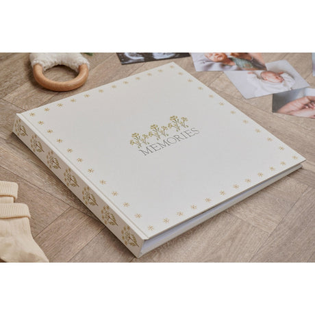 Square Memories linen photo album in cream, with a gold floral border, gold floral sprigs and grey lettering on the cover, shown on a wooden floor beside baby photos and keepsakes.