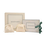 Square cream keepsake box labelled "KEEPSAKES" with floral-bordered lid, paired with a "MEMORIES" box and two cotton drawstring pouches, finished with a green ribbon tie, on a plain white background.