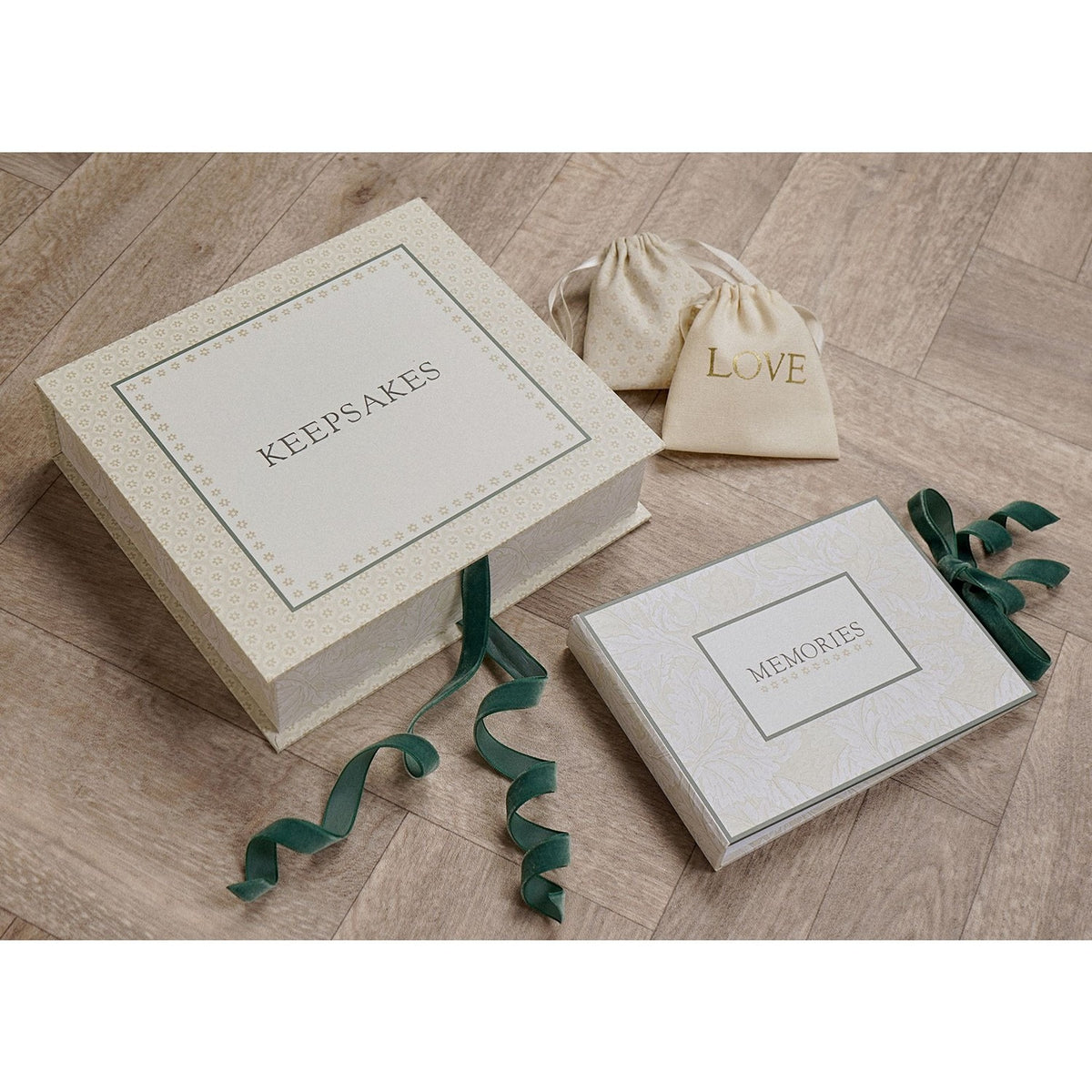 Square cream keepsake box with "KEEPSAKES" lettering and floral border, tied with a green ribbon, shown beside a matching "MEMORIES" photo album and two drawstring pouches on a wooden floor.