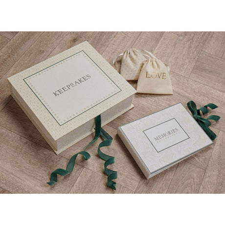 Square cream keepsake box with "KEEPSAKES" lettering and floral border, tied with a green ribbon, shown beside a matching "MEMORIES" photo album and two drawstring pouches on a wooden floor.