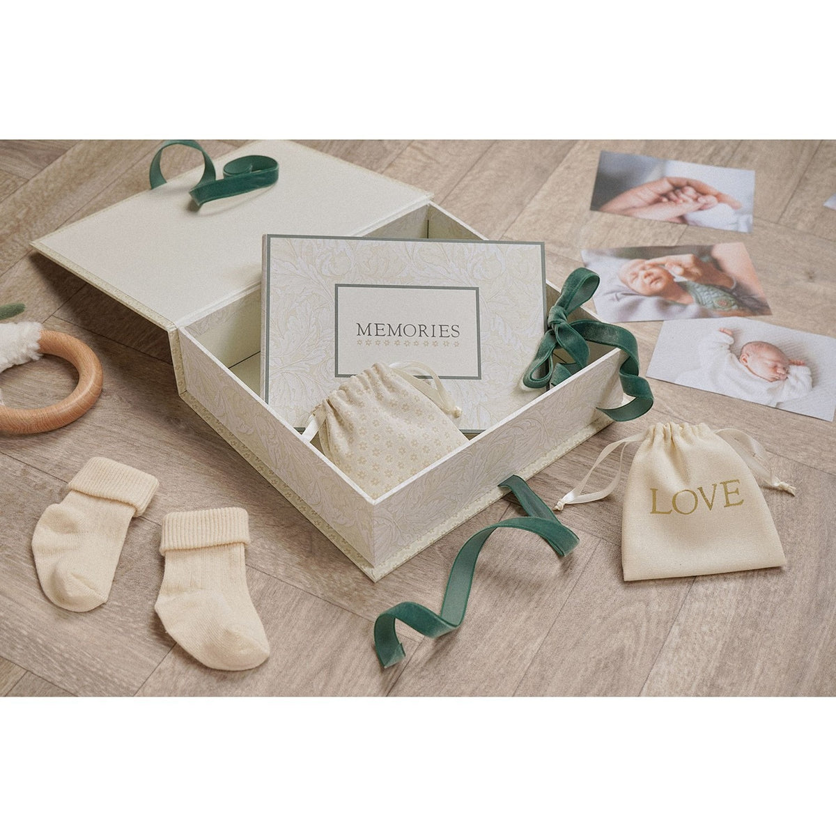 Square cream baby keepsake box with green ribbon tie, open to show a Memories card and drawstring pouches, displayed with baby socks, photos and a teething ring.