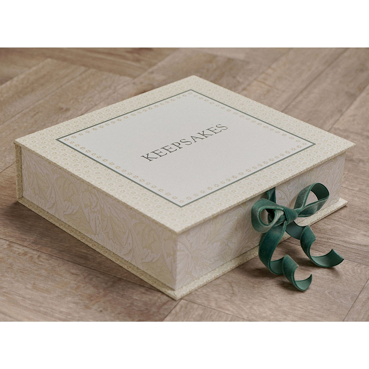 Square cream keepsake box with "KEEPSAKES" printed on the lid, floral border, leafy side pattern, and a green ribbon tied in a bow at the front, shown on a wooden floor.