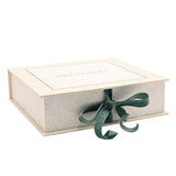 Square cream keepsake box with "KEEPSAKES" printed on the lid, floral border, leafy patterned sides, and a green ribbon tied in a bow at the front, shown on a plain white background.