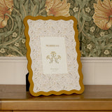 Rectangular metal photo frame with wavy ochre border and bird and floral patterned mount, holding a 7 x 5in print, displayed on a wooden sideboard against floral wallpaper.