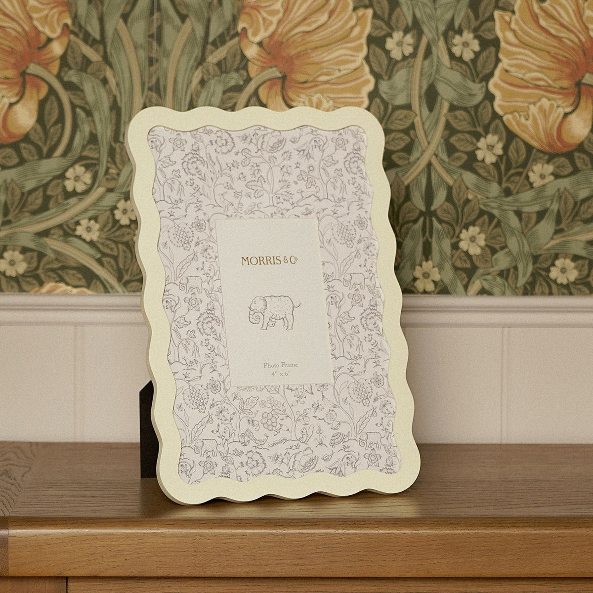 Cream metal photo frame with scalloped wavy border, featuring a botanical mount printed with grey floral and animal motifs, displayed against a leafy patterned wallpaper.