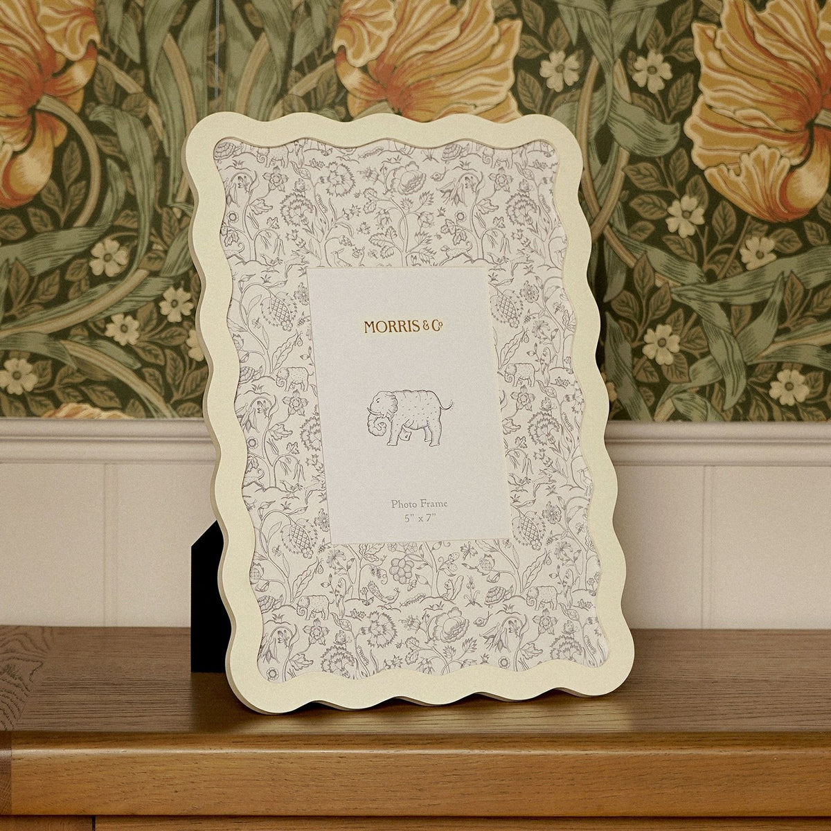 Baby Wiggle Metal Photo Frame in cream with a wavy scalloped border and botanical-print mount, displayed on a wooden sideboard against floral wallpaper.
