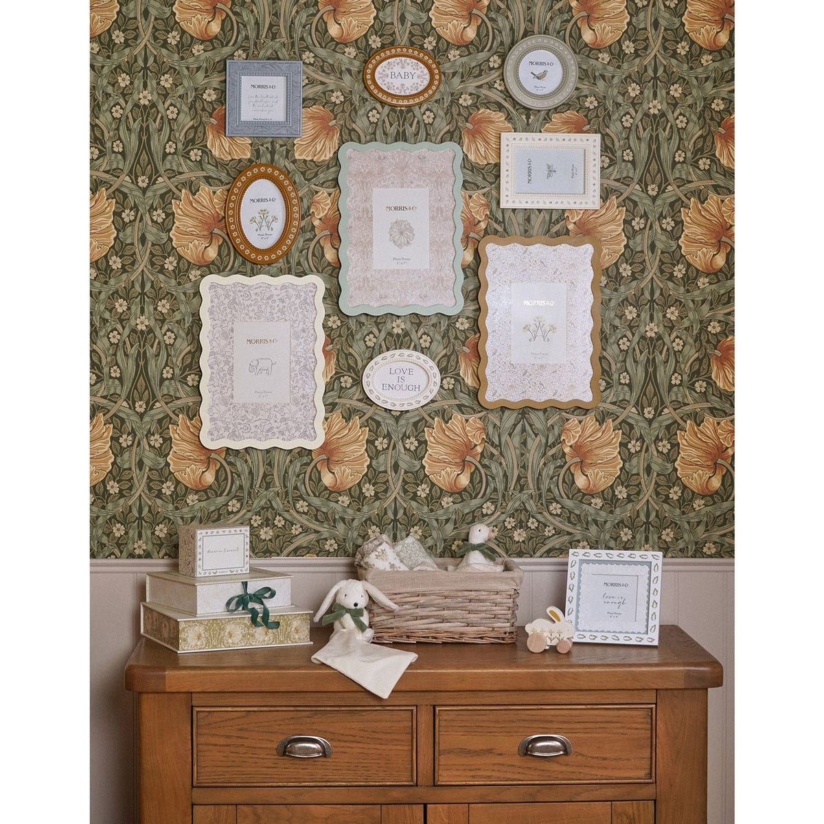 Cream metal photo frame with wavy scalloped border displayed on a wooden dresser, part of a gallery wall of frames on floral wallpaper in a nursery setting.