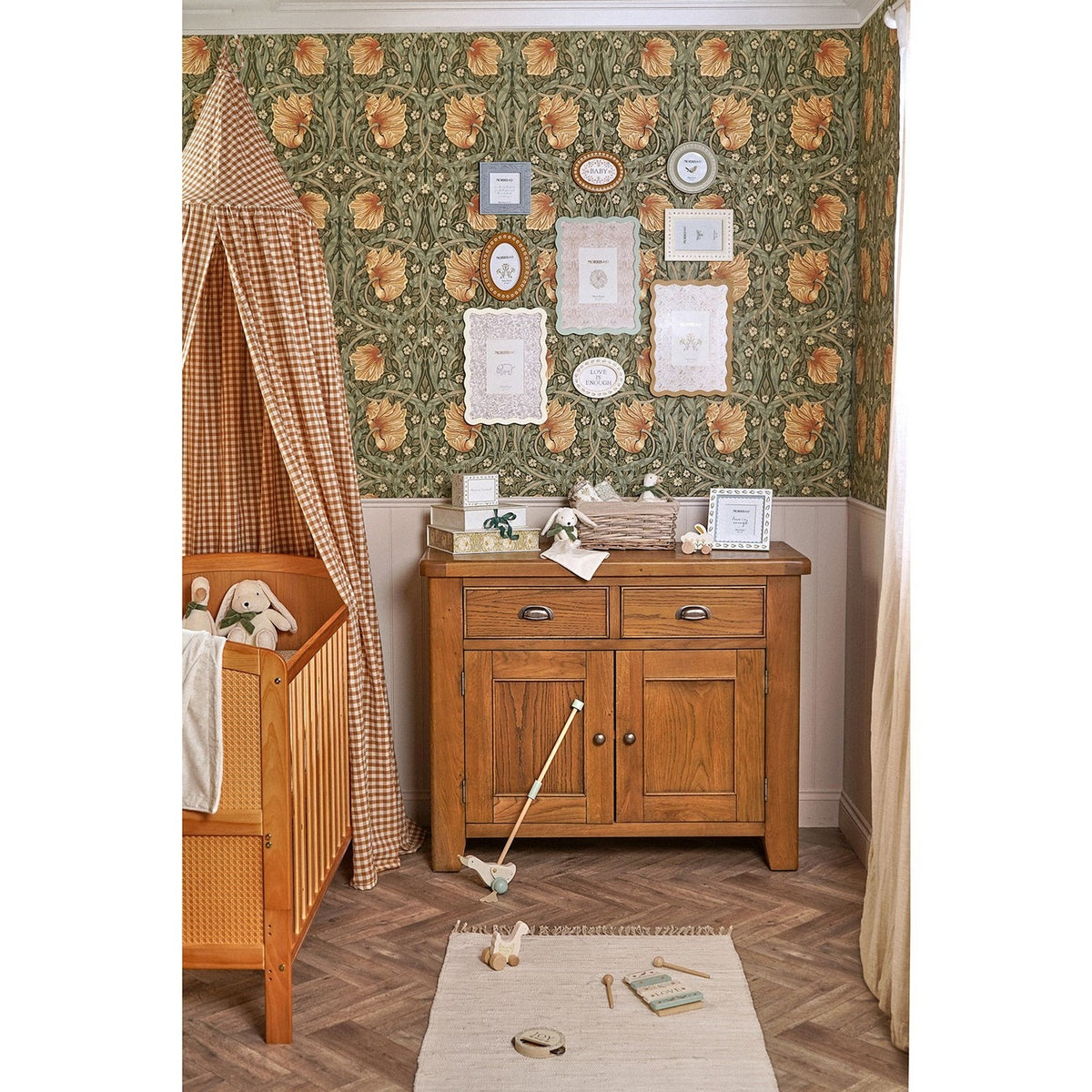 Cream metal photo frame with wavy scalloped border and botanical-print mount, displayed on a wooden sideboard in a nursery setting.