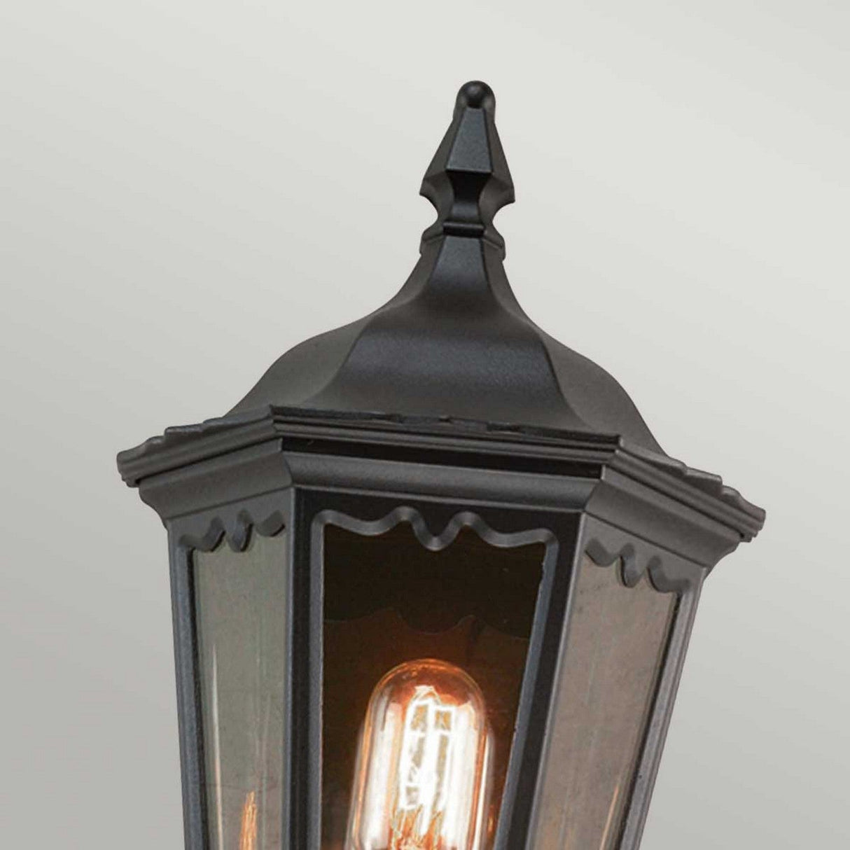 Close-up of the Medstead 1 Light Outdoor Wall Light With PIR in black, featuring a pointed metal top and clear glass panels showcasing an exposed glowing filament bulb inside, all set against a plain light gray backdrop. The timeless design highlights the elegance of its black finish.