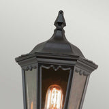 Close-up of the Medstead 1 Light Outdoor Wall Light With PIR in black, featuring a pointed metal top and clear glass panels showcasing an exposed glowing filament bulb inside, all set against a plain light gray backdrop. The timeless design highlights the elegance of its black finish.