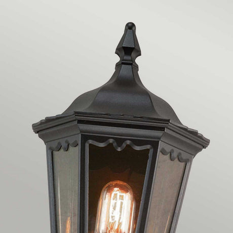 Close-up of the Medstead 1 Light Outdoor Wall Light With PIR in black, featuring a pointed metal top and clear glass panels showcasing an exposed glowing filament bulb inside, all set against a plain light gray backdrop. The timeless design highlights the elegance of its black finish.