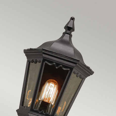 The Medstead 1 Light Outdoor Wall Light With PIR in black features a vintage design with a pointed top and clear glass panels that highlight an illuminated filament bulb. With an IP44 rating, it is ideal for complementing a neutral gray background and enhancing any exterior setting.