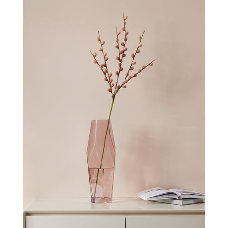Mehakit artificial blossom branch in pink, displayed in a faceted pink glass vase on a white cabinet against a muted pink wall, with an open book nearby.