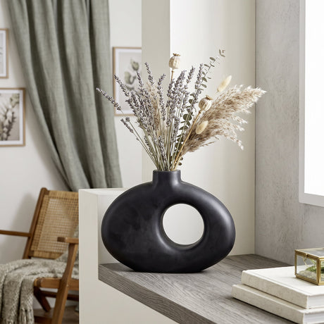 Melobody mango wood vase in matt black with a flat oval body, off-centre round cutout and short neck, styled with dried florals on a shelf.