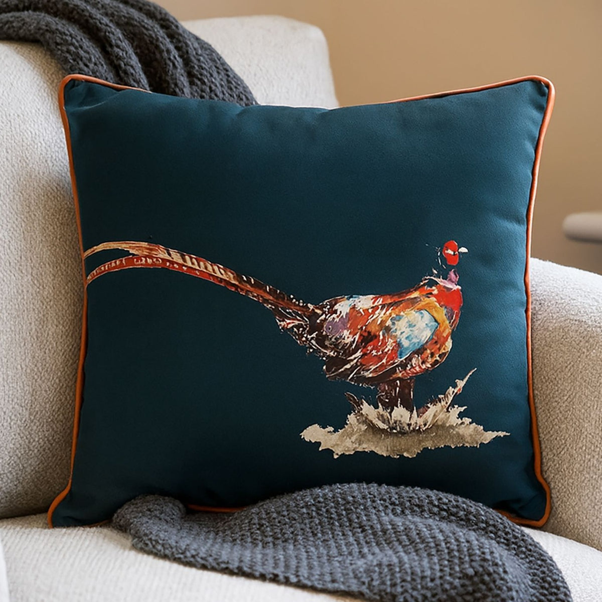 Square pheasant cushion in deep teal blue with orange piping, featuring a painterly russet and copper pheasant print, shown on a cream armchair with a grey knit throw.