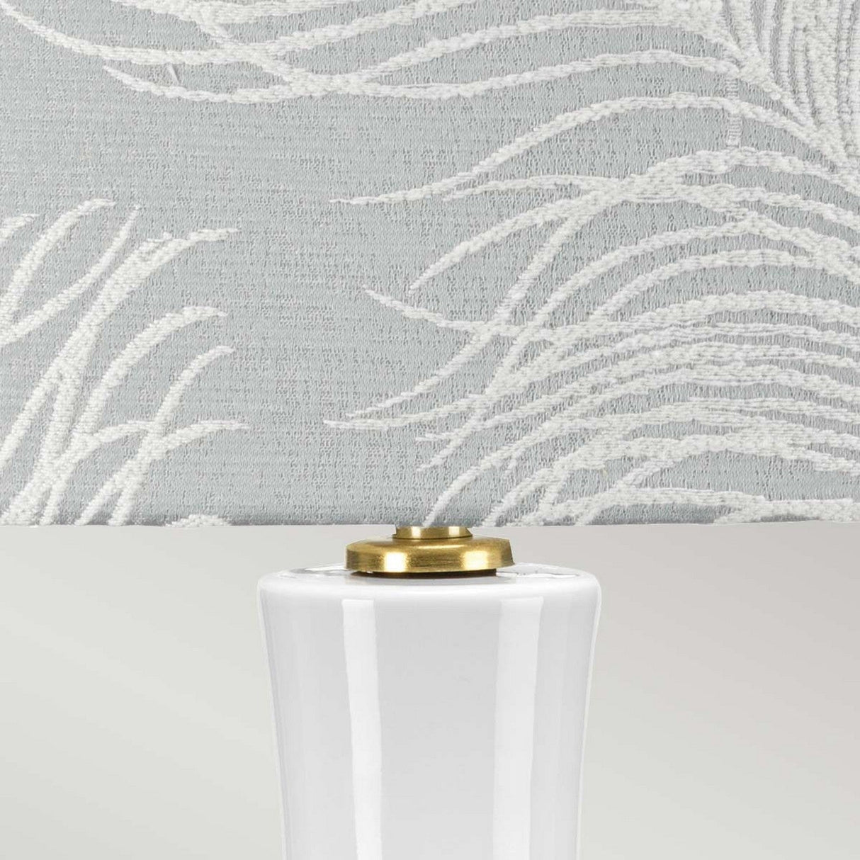Take a closer look at the Milo 1 Light Table Lamp, featuring a white ceramic base paired with an aged brass lamp holder. The light gray lampshade is beautifully adorned with a textured, embroidered pattern that resembles abstract fern leaves.