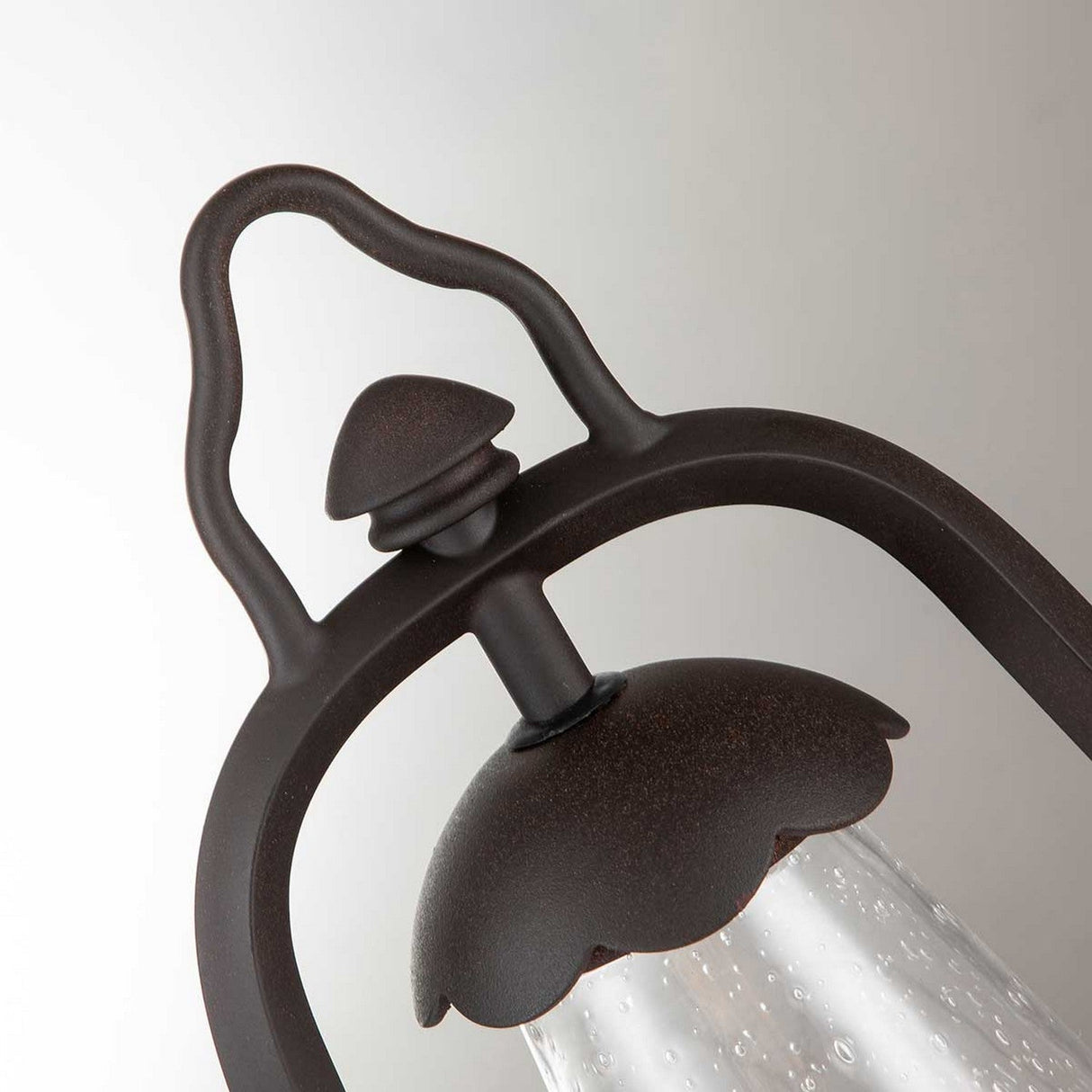 Close-up of the Miners 1 Light Pedestal Light in Old Bronze, showcasing its dark metal finish. The design includes a handle and scalloped edges surrounding textured, bubbled glass. This outdoor light is set against a neutral, light gray backdrop.