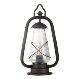 The Miners 1 Light Pedestal Light in Old Bronze features a rustic black frame with a glass enclosure and a metal wire cross design around the center, reminiscent of antique lanterns. Its vintage-style loop handle at the top enhances its antique appearance, making it ideal for use as an outdoor light.