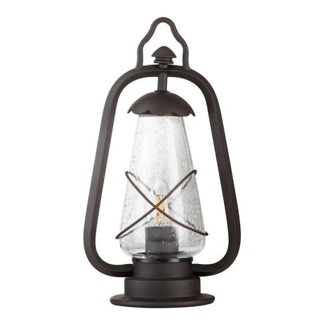 The Miners 1 Light Pedestal Light in Old Bronze features a rustic black frame with a glass enclosure and a metal wire cross design around the center, reminiscent of antique lanterns. Its vintage-style loop handle at the top enhances its antique appearance, making it ideal for use as an outdoor light.