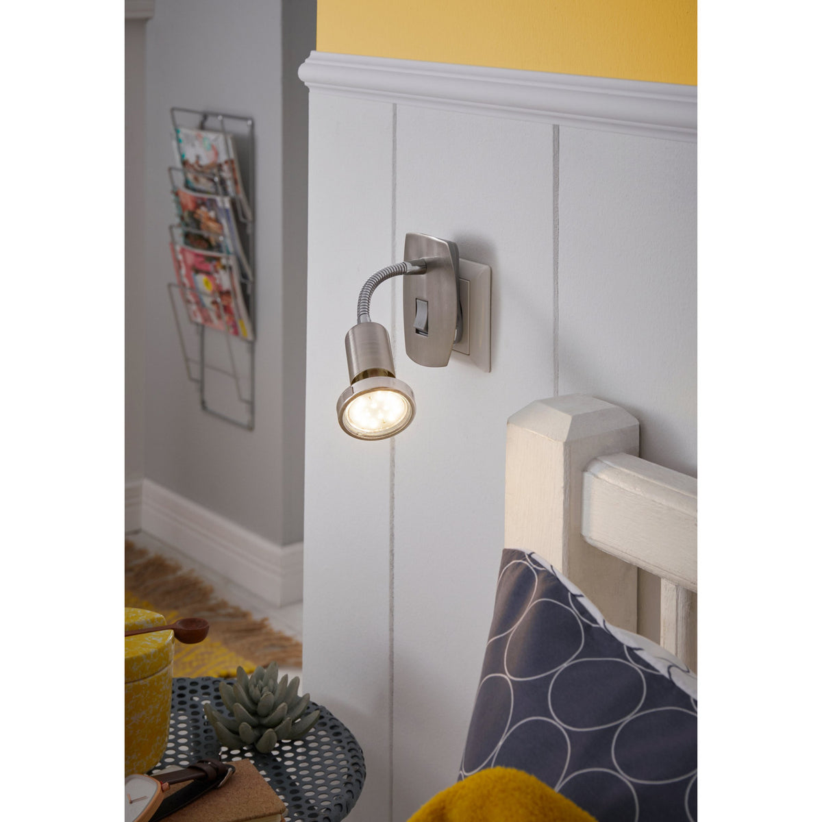 Mini 4 plug-in spotlight in satin nickel and chrome with gooseneck arm and GU10 bulb, shown mounted above a bedroom headboard with warm white light on.
