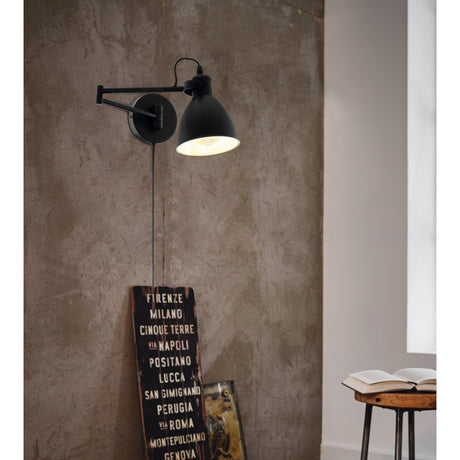 Minvora Wall Light in black steel with hinged swing arm and bell-shaped shade, shown lit on a textured wall above a vintage sign and stool.