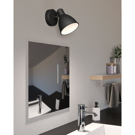 Minvora black wall light with bell-shaped steel shade, white interior, and round backplate, mounted above a wall mirror in a bathroom setting.
