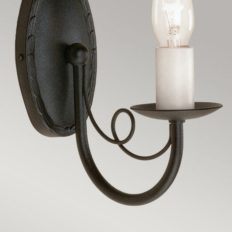 A close-up of the Minster 1 Light Wall Light - Black highlights its vintage craftsmanship, featuring a black metal arm and decorative loop. The wall sconce displays a bare bulb on a white candle-like socket against a neutral light gray backdrop.