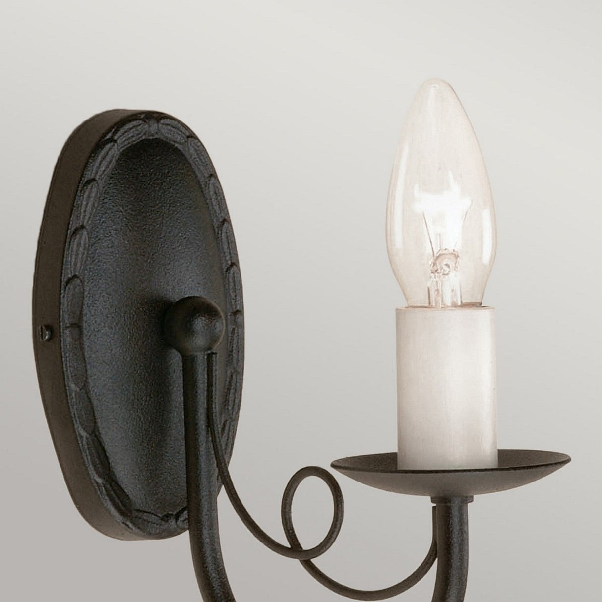 The Minster 1 Light Wall Light - Black is a wall-mounted sconce with a black finish and a candle-shaped light bulb. It features a round backplate and an elegant design accented by curled metalwork, showcasing traditional craftsmanship that adds timeless charm to any neutral background.