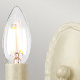 A close-up of the Minster 1 Light Wall Light – Ivory/Gold Patina, featuring a glowing candle-shaped bulb with visible filaments, casts a warm glow beside a decorative, textured ceramic surface. The light's gentle illumination contrasts beautifully against the ivory background, adding depth and elegance to the scene.