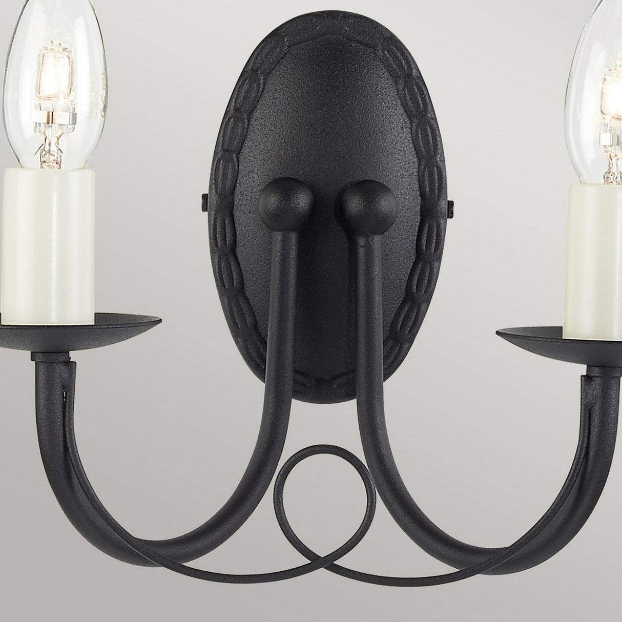 The Minster 2 Light Wall Light - Black is an elegant fixture designed with two curved arms, each supporting a candle-style light bulb. It is mounted on an oval backplate and seamlessly blends into both modern and traditional interiors, casting a warm glow when the bulbs are turned on.